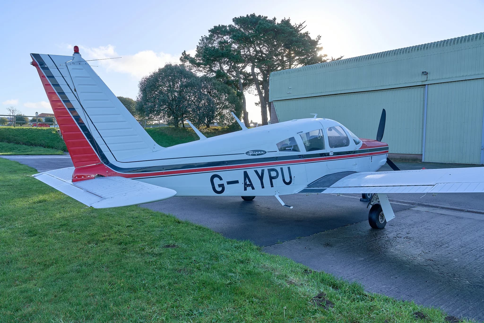 A rear-side view of the white and red Piper Cherokee G-AYPU parked near a hangar and trees.