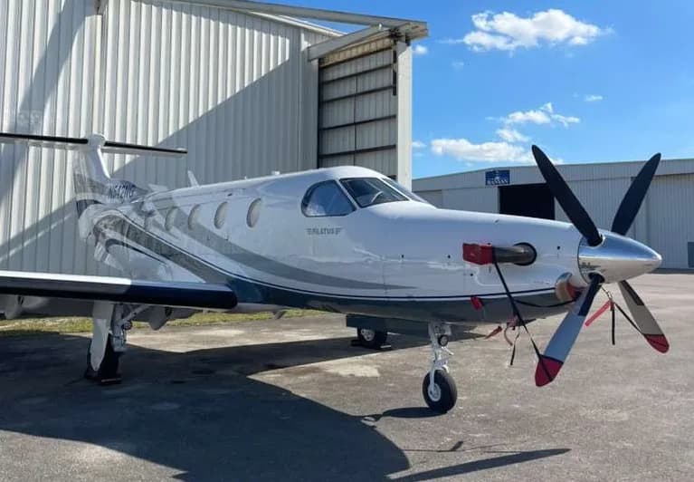 A white Pilatus PC-12 NG aircraft with blue and grey stripes, registration N542NG, parked in front of a hangar.