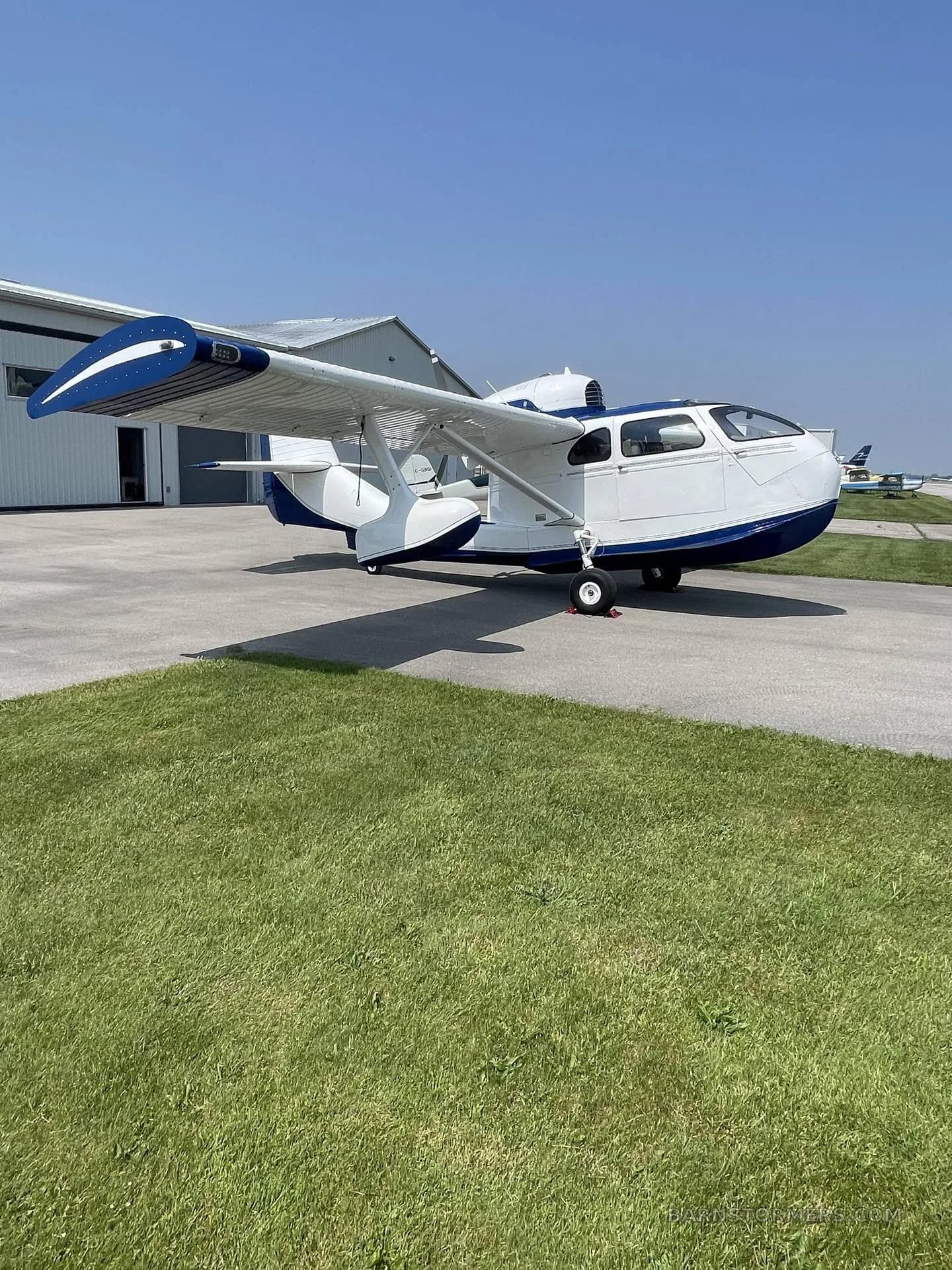 A white and blue amphibious aircraft with its wings folded, parked on an airfield tarmac next to a hangar.