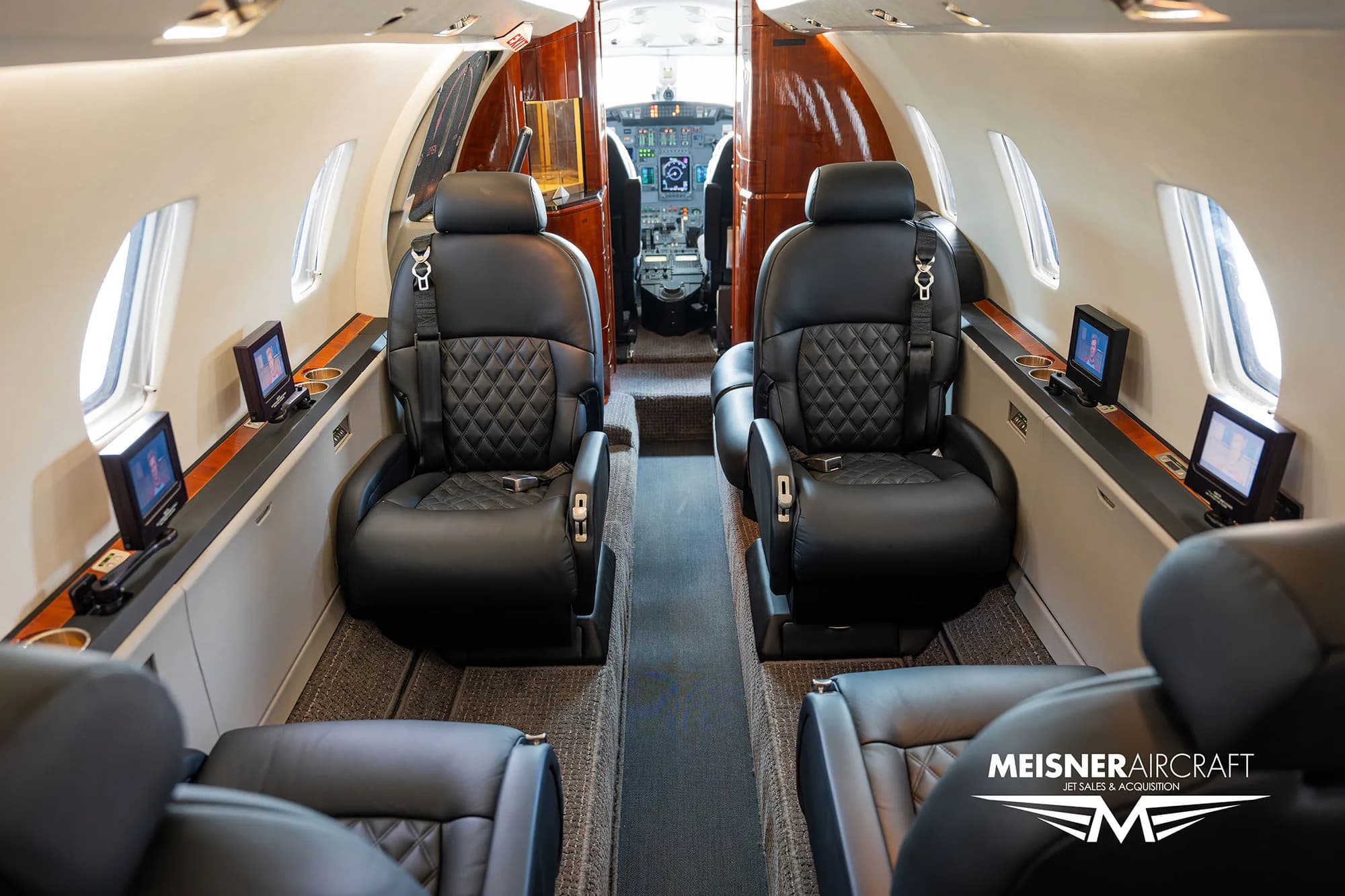 The luxurious interior cabin of a Citation Excel business jet with black leather seats and wood paneling, looking towards the cockpit.