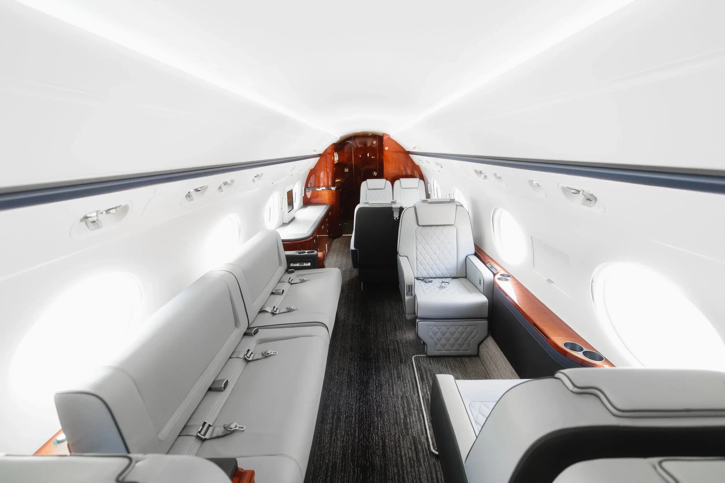 A wide view of the private jet's cabin, showing a long light gray couch on one side and individual seats on the other.