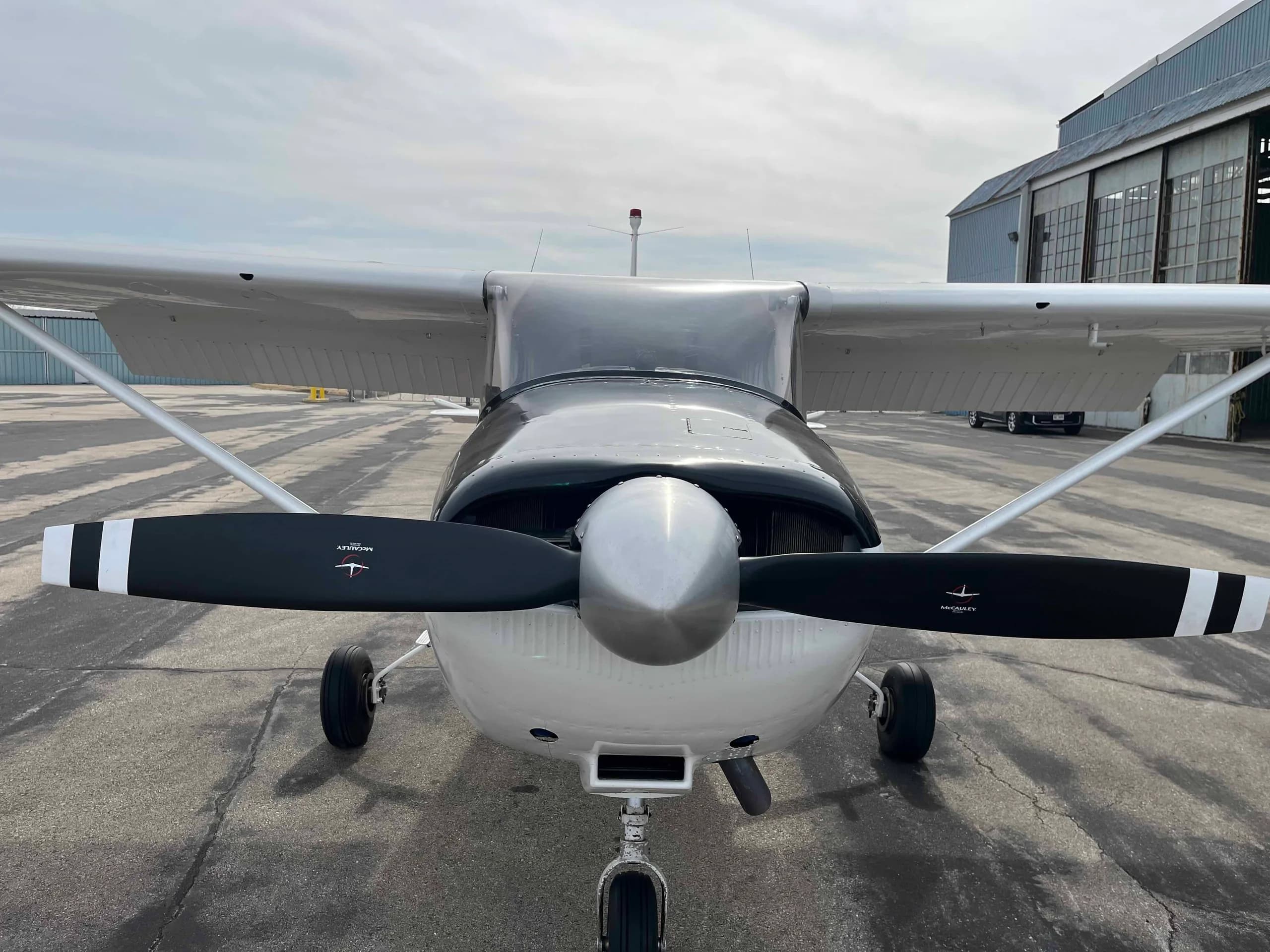A head-on view of a white Cessna 182 aircraft with a black propeller and nose cone, parked on an asphalt apron.