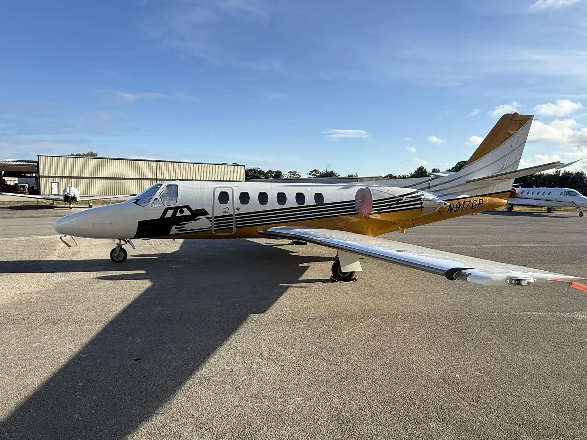 A Cessna Citation jet, N917GP, with white and gold paint scheme, parked on an airfield with hangars in the background.