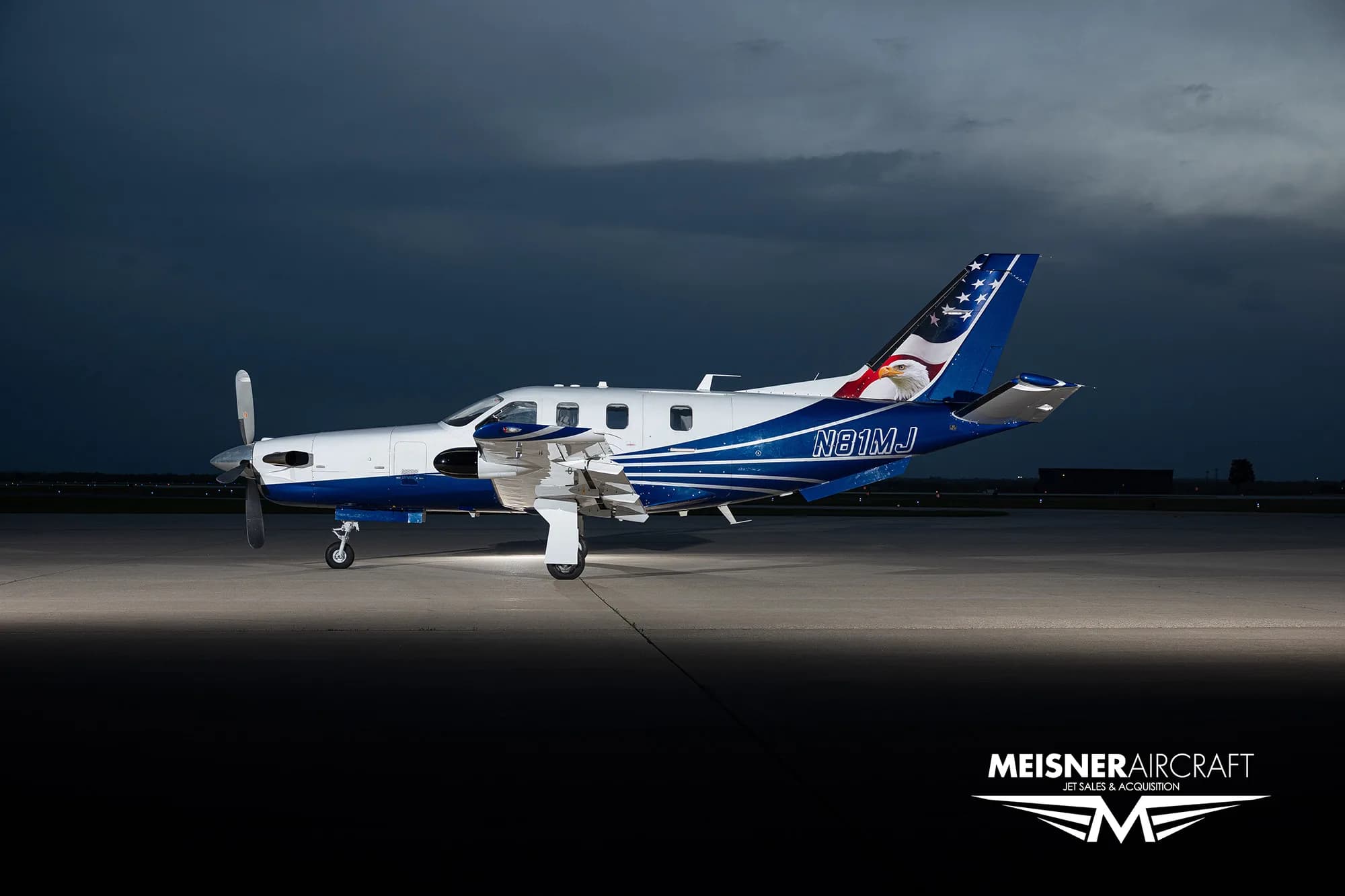 A TBM turboprop aircraft with a blue and white paint scheme featuring an eagle on the tail, parked on an airfield at dusk.
