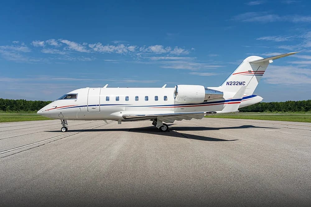 A white Bombardier Challenger business jet with red and blue stripes is parked on an asphalt runway under a clear blue sky.