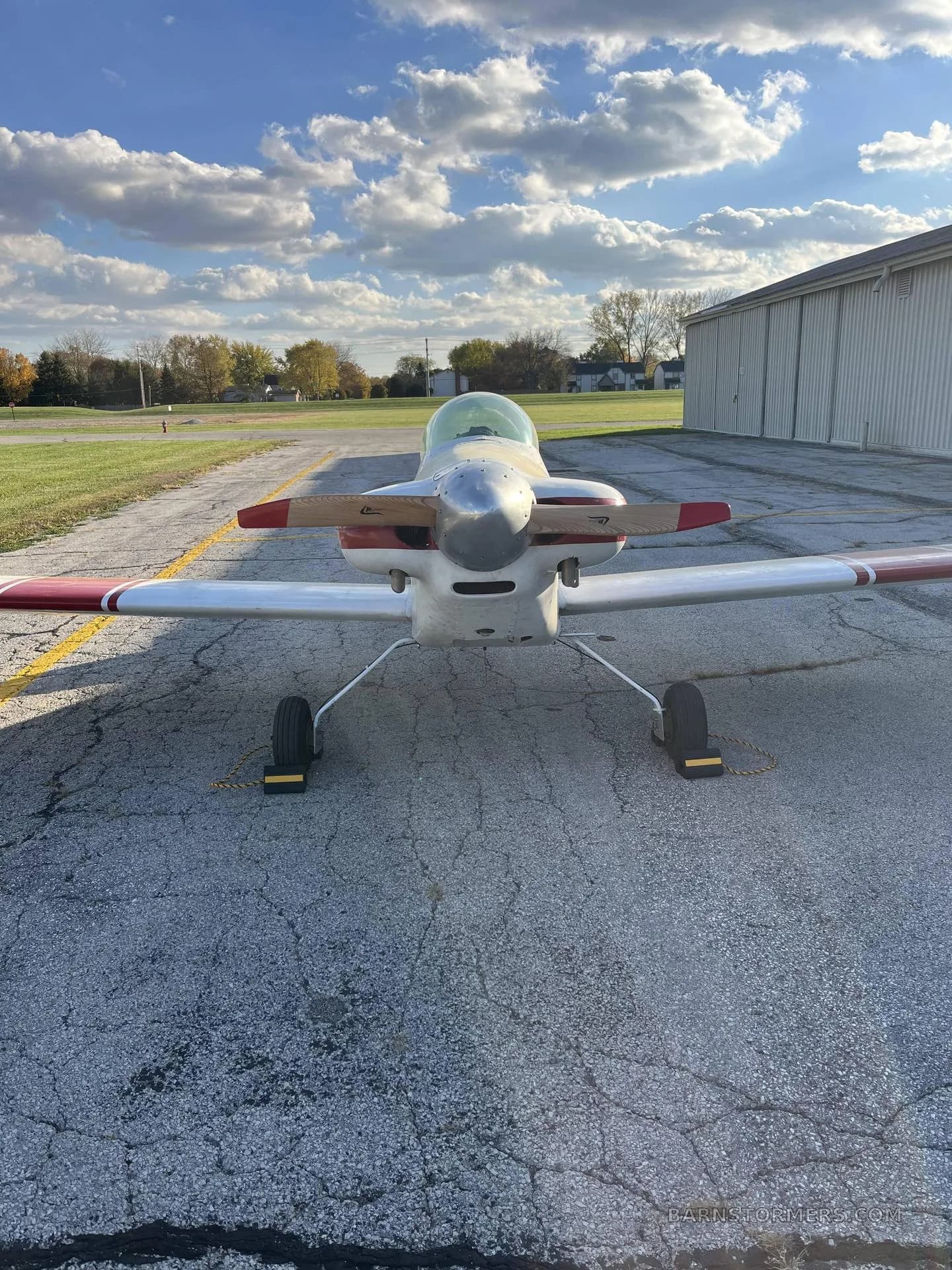 A front view of a white and red single-engine aircraft with a wooden propeller, parked on an asphalt apron in front of a hangar.