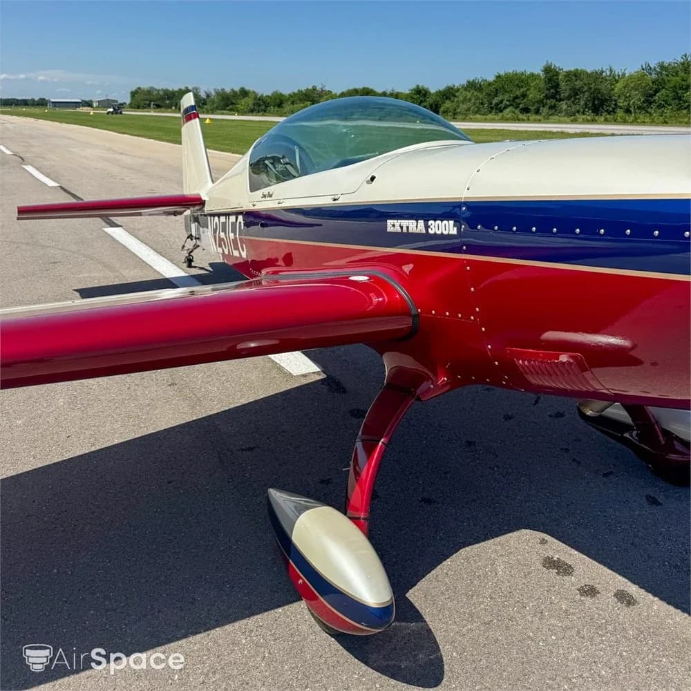 A close-up view of the front fuselage and wing of the red, white, and blue Extra 300L aircraft, N251EC, on an airfield.