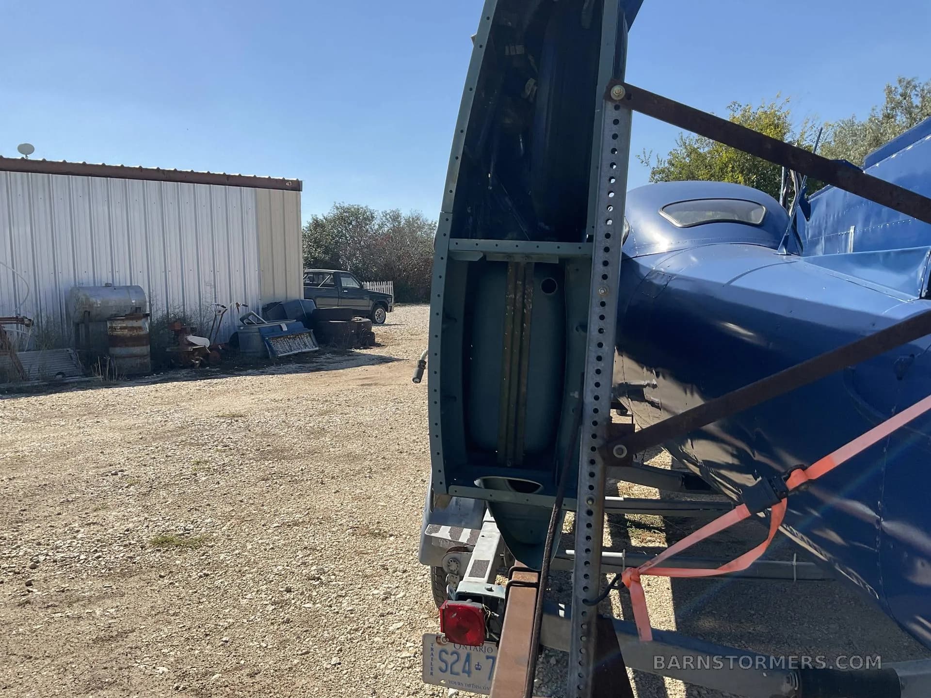 A close-up of the rear section of a blue aircraft fuselage on a trailer, showing internal structure and a fuel tank, with a white building and junk in the background.