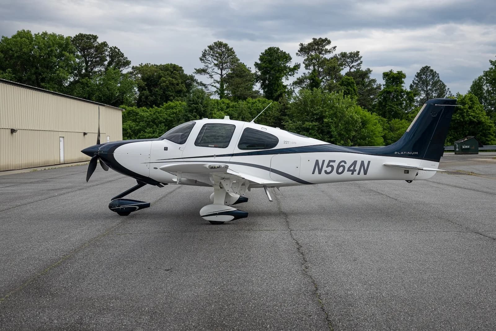 A white and dark blue Cirrus SR22T aircraft with registration N564N parked on an asphalt ramp with trees in the background.
