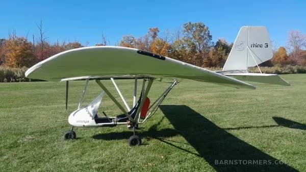 A white ultralight aircraft with 'prototype' on its nose and 'rhine 3' on its tail is parked on a grassy field under a clear blue sky.
