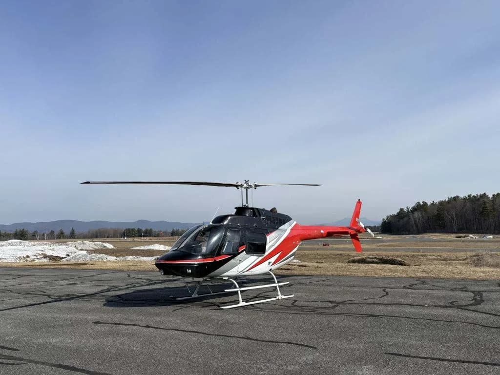 A red, black, and white helicopter with skids on an airfield with mountains in the background.