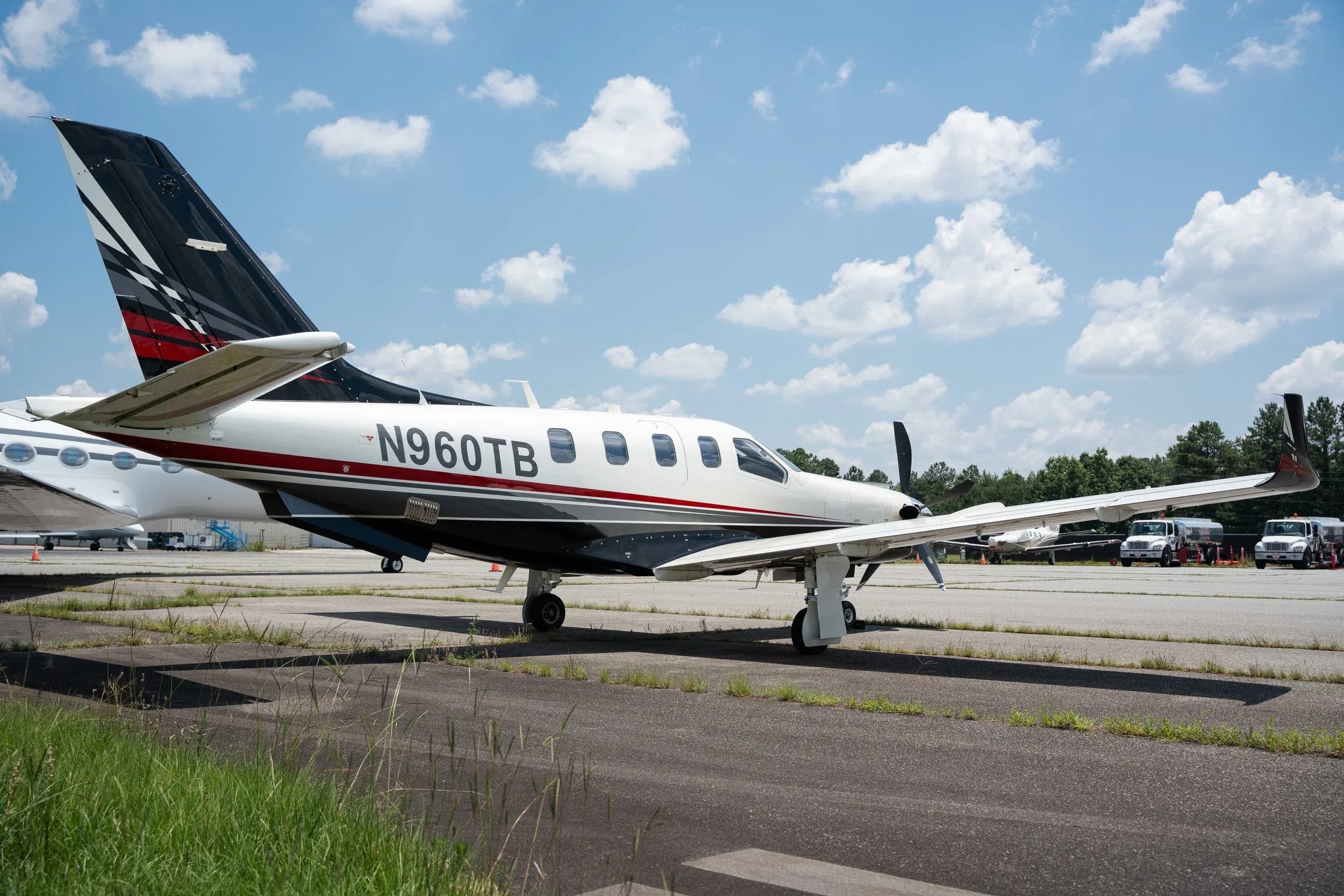 A side view of a white and black TBM 960 aircraft with registration N960TB, parked on an airport tarmac with other aircraft in the background.