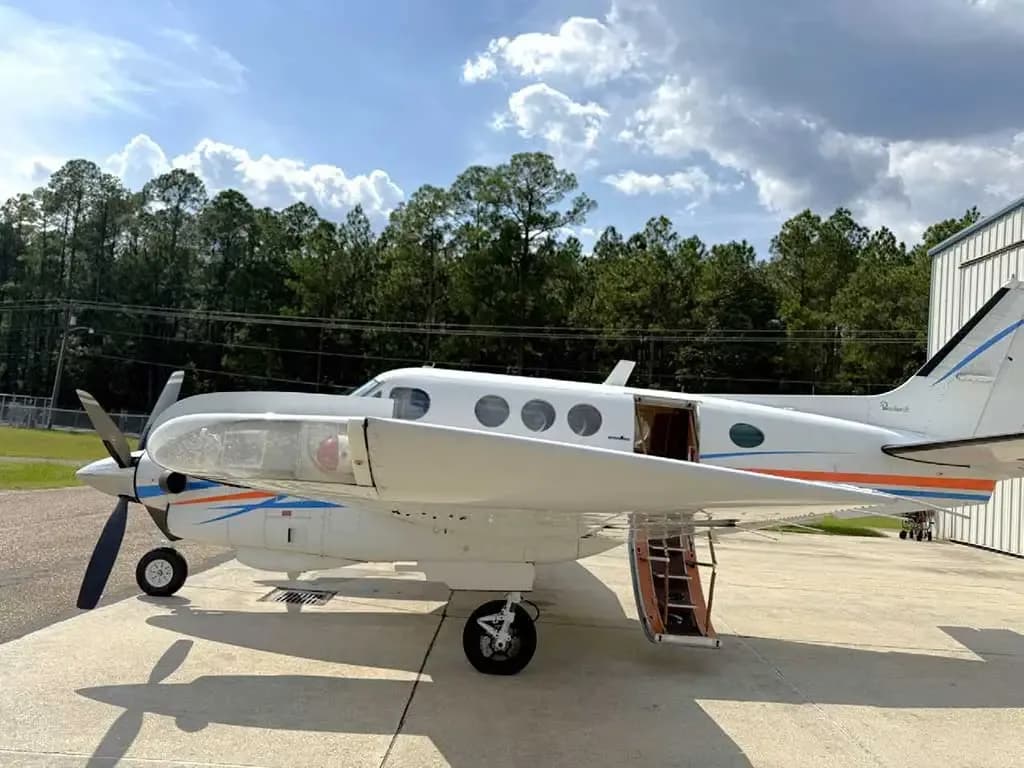 A white twin-engine turboprop aircraft with blue and orange stripes, parked on a tarmac next to a hangar with its passenger door open.