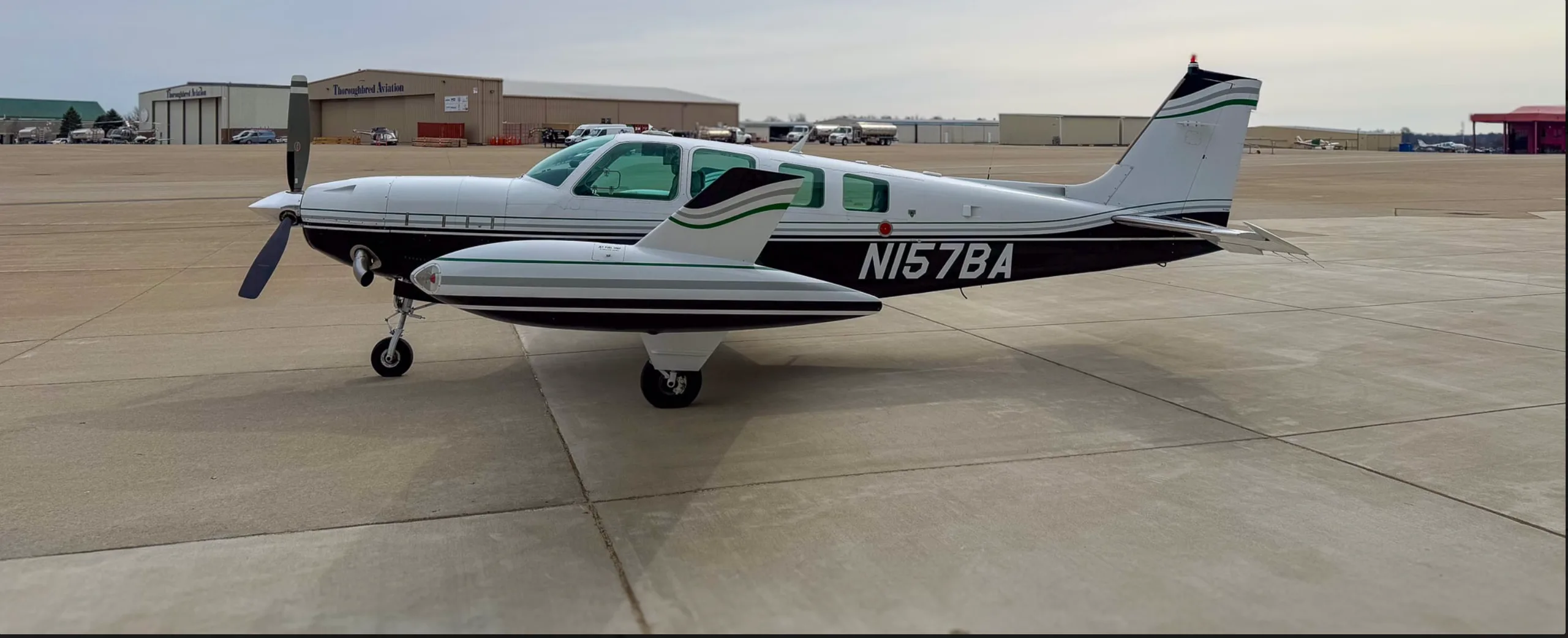 A side view of the white and black Beechcraft Baron N157BA parked on a tarmac with airport buildings in the background.
