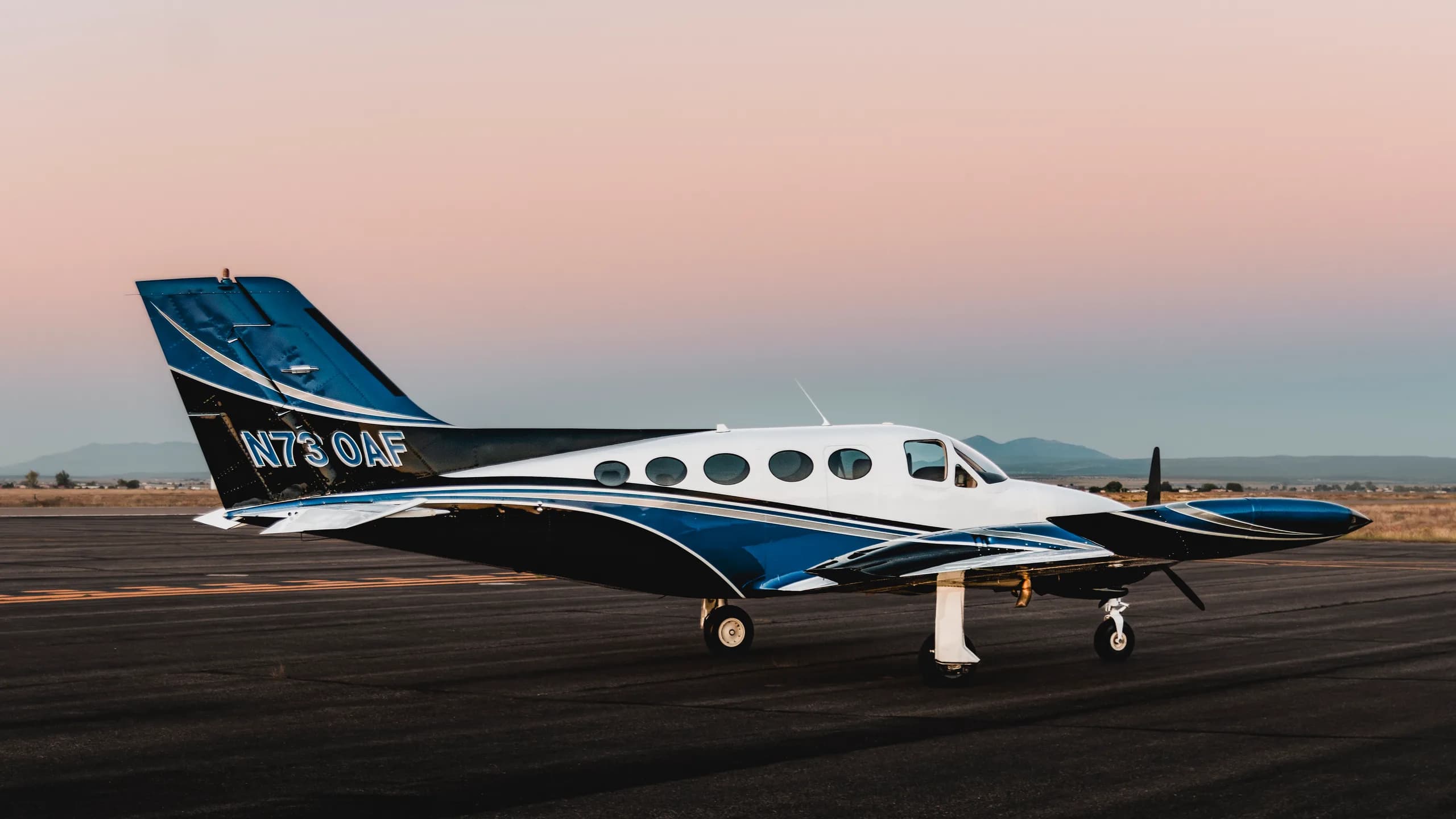 A white and blue twin-engine aircraft parked on a runway at sunset.