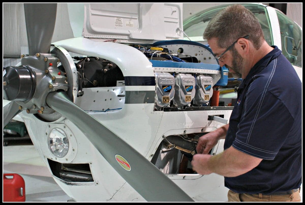 A mechanic in a hangar working on the open engine compartment of a white propeller aircraft.