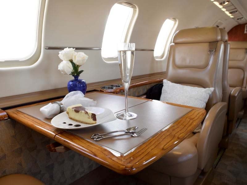 A view inside the aircraft cabin, showing a table set with cheesecake, champagne, and white roses next to a tan leather seat.