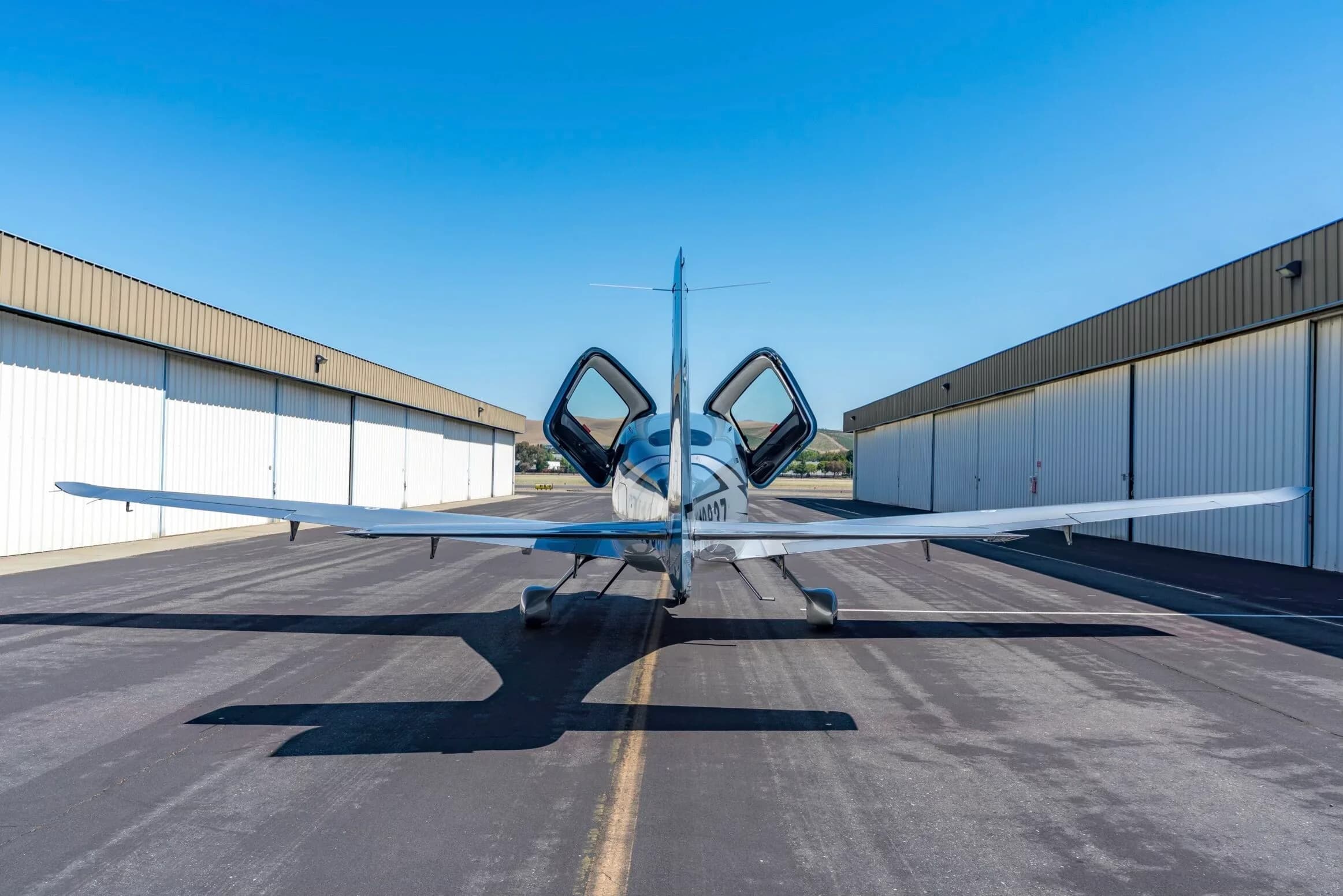 Rear view of a light blue and silver Cirrus SR22T aircraft, N1383Z, with its doors open, parked between hangars.