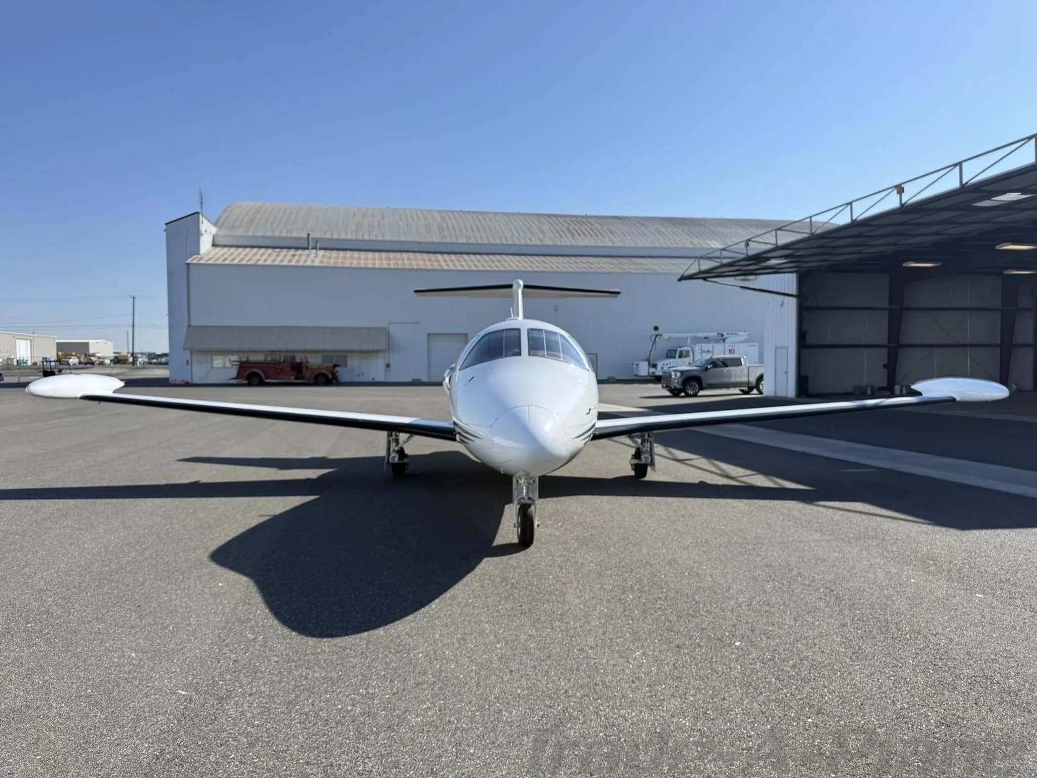 Front view of a white private jet parked on an asphalt apron in front of a hangar under a clear sky.