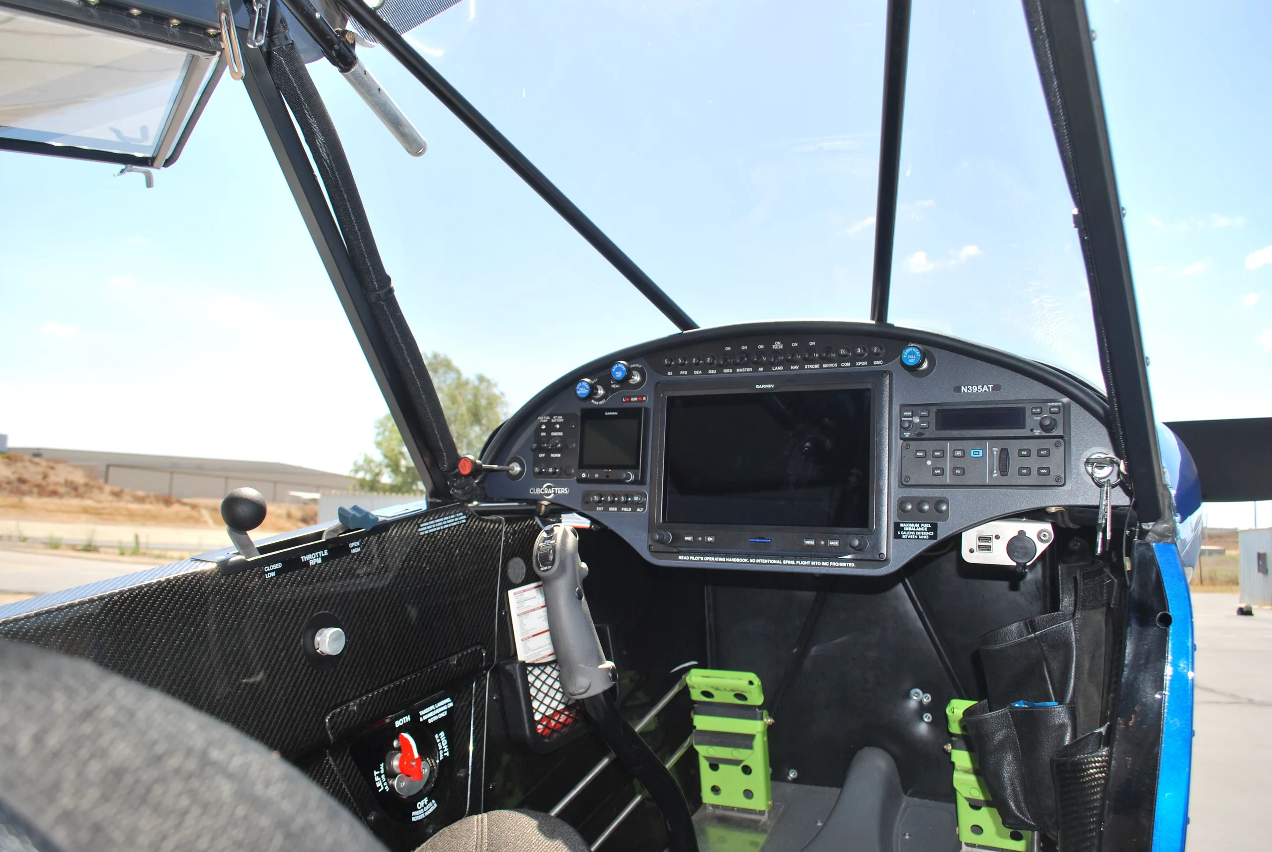 A detailed view of the cockpit of a Carbon Cub FX3, showing the instrument panel with a large Garmin display and green rudder pedals.