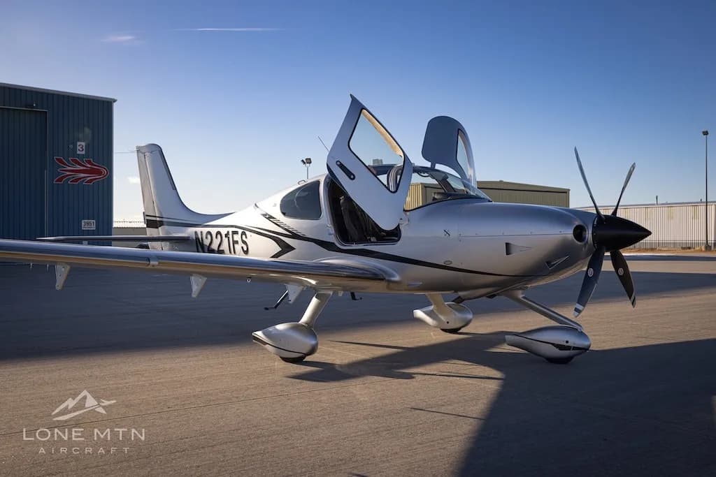 A three-quarter front view of the silver Cirrus SR22T aircraft N221FS with its doors open, parked on an airfield.