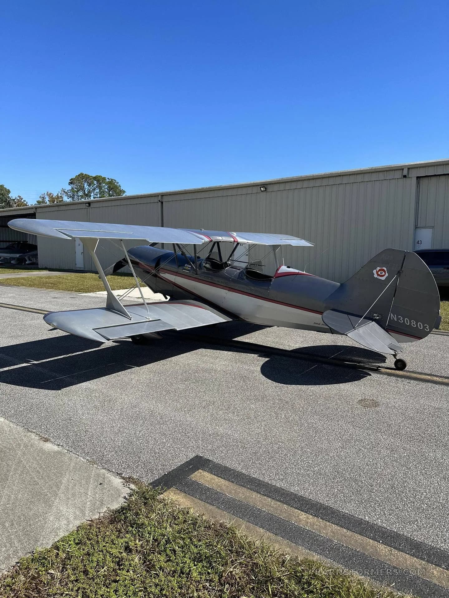 A gray and white biplane with red accents is parked on an asphalt tarmac next to a large metal hangar.