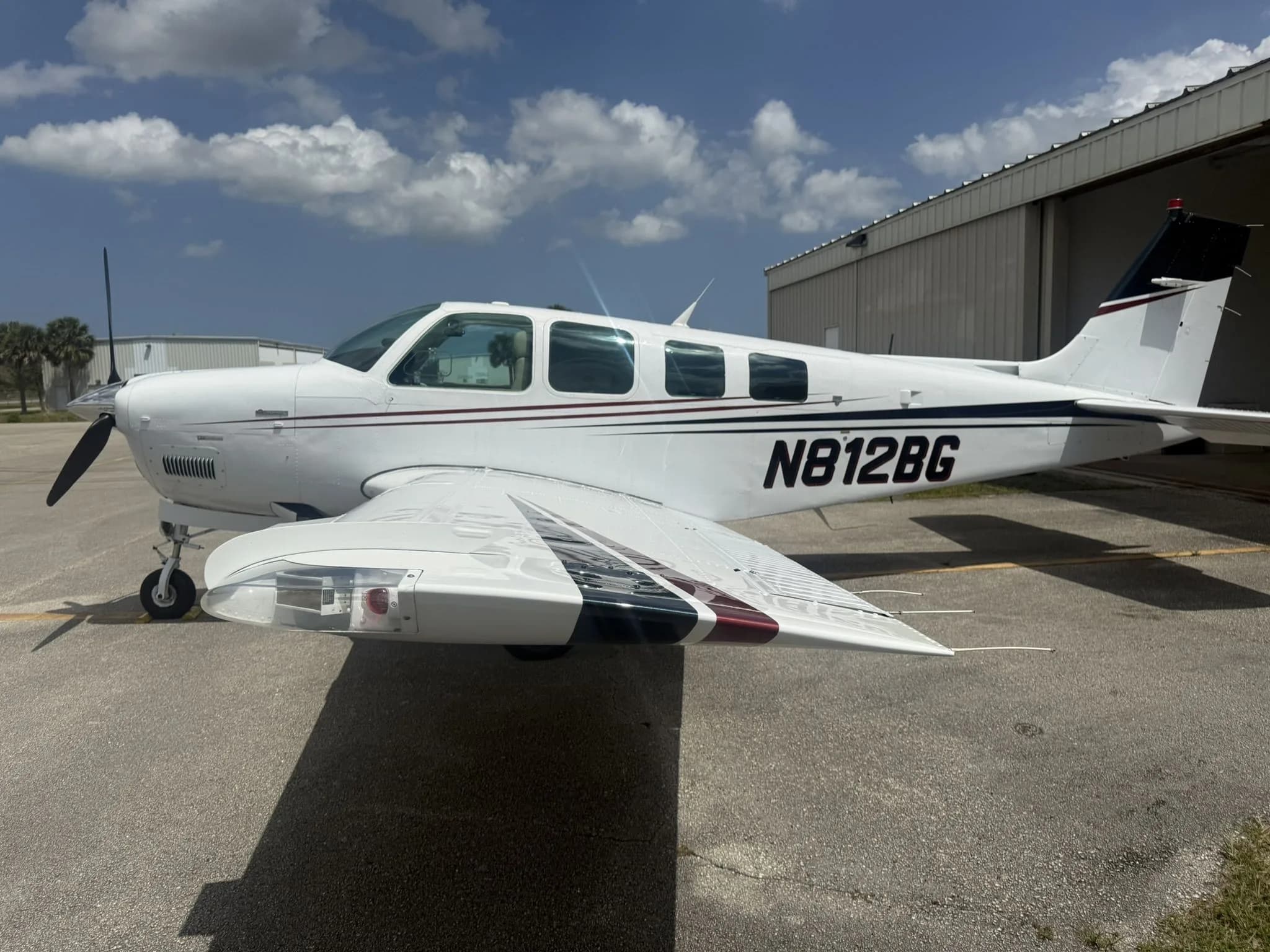 Side view of a white Beechcraft Bonanza N812BG with dark red and black stripes and wing details on an airfield.