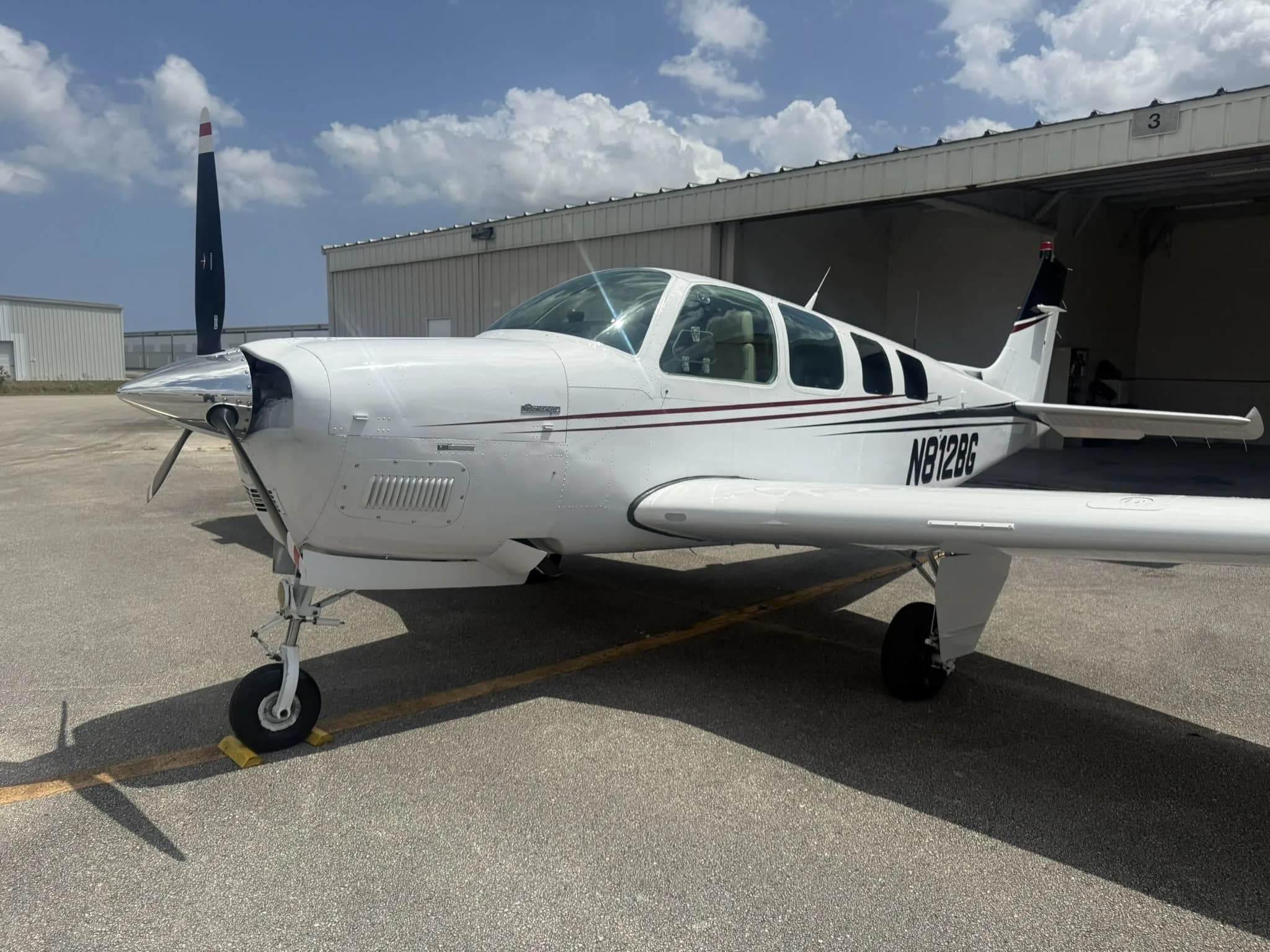 Front three-quarter view of a white Beechcraft Bonanza N812BG with dark red and black stripes on an airfield.