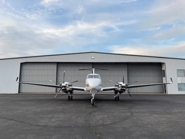 A white Beechcraft King Air with red and black stripes parked on an tarmac in front of a hangar, viewed head-on.
