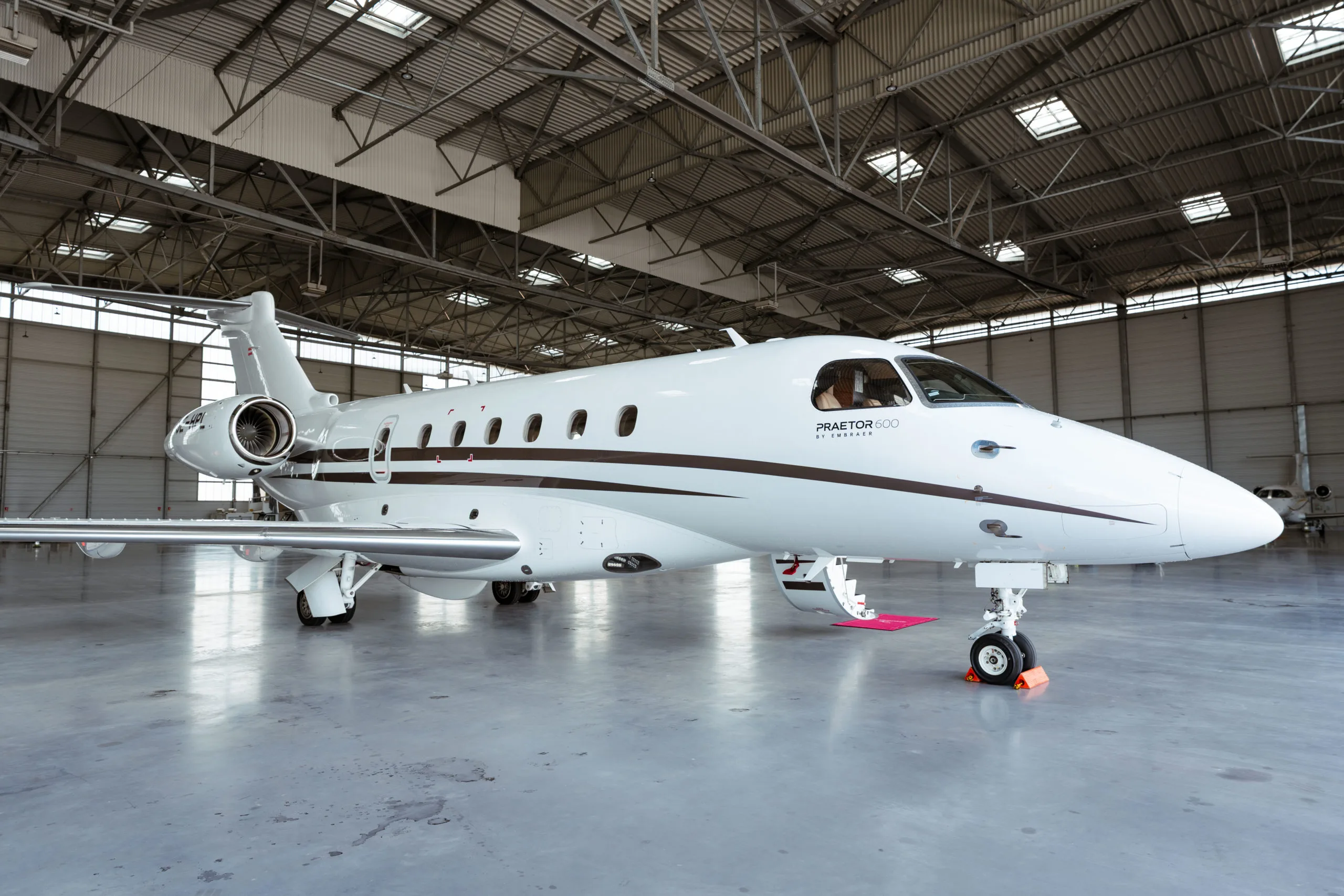 A white Embraer Praetor 600 business jet with a brown stripe, parked inside a large hangar.