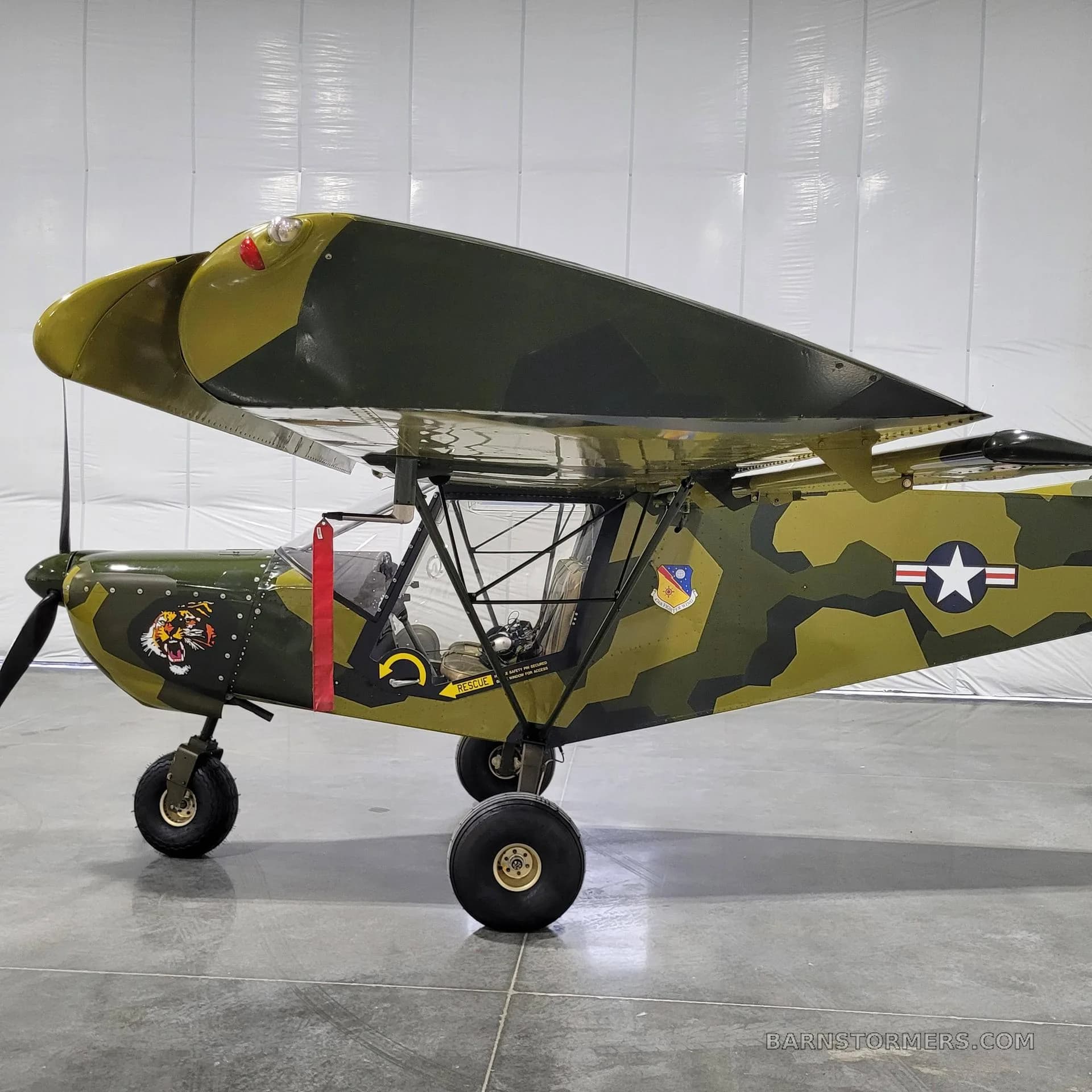 A side view of a camouflage-painted, high-wing aircraft featuring a tiger decal and a US Air Force star insignia, parked in a hangar.
