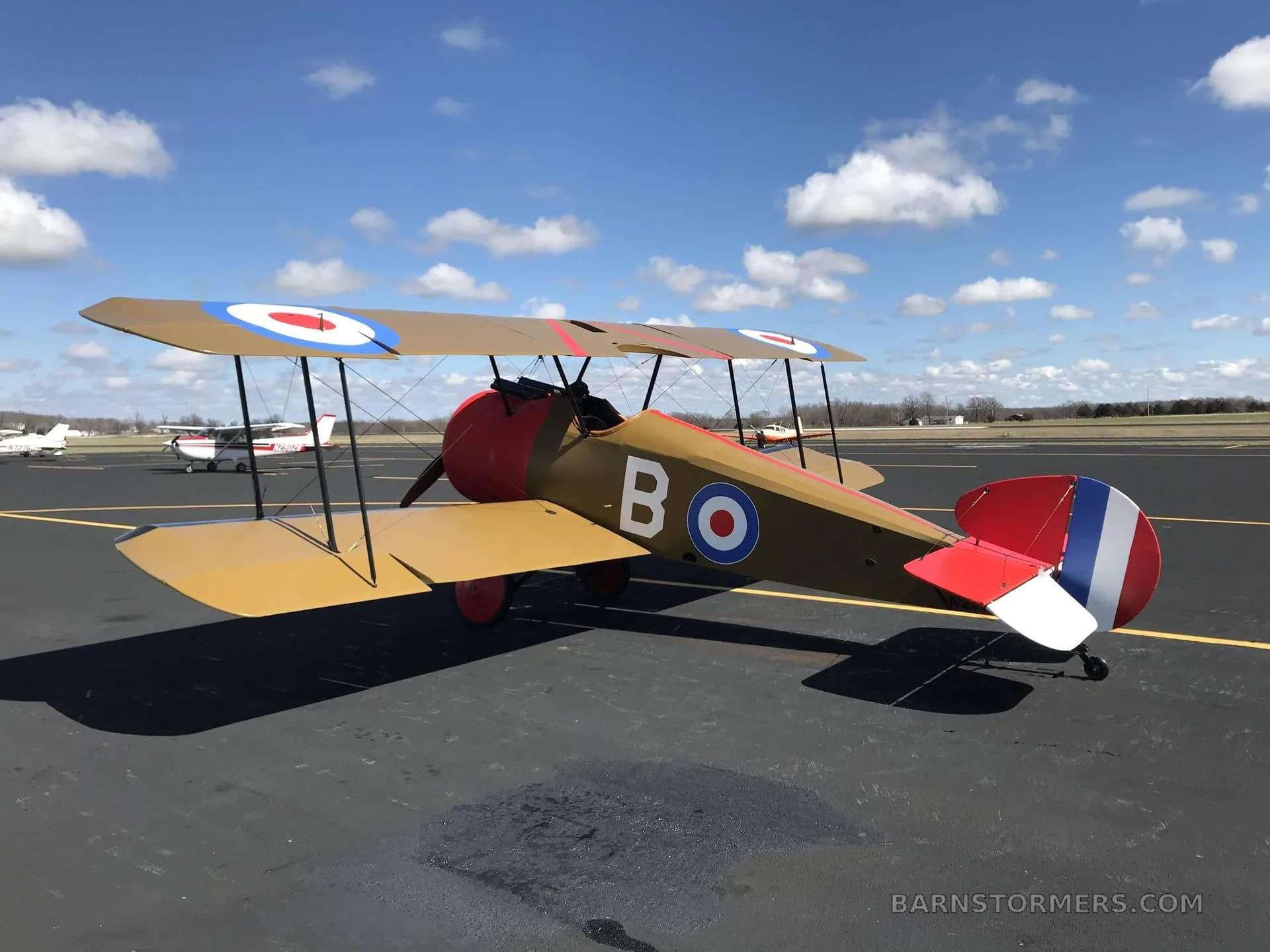 A tan and red biplane with British roundels is parked on an airport tarmac with other small planes and hangars in the background.