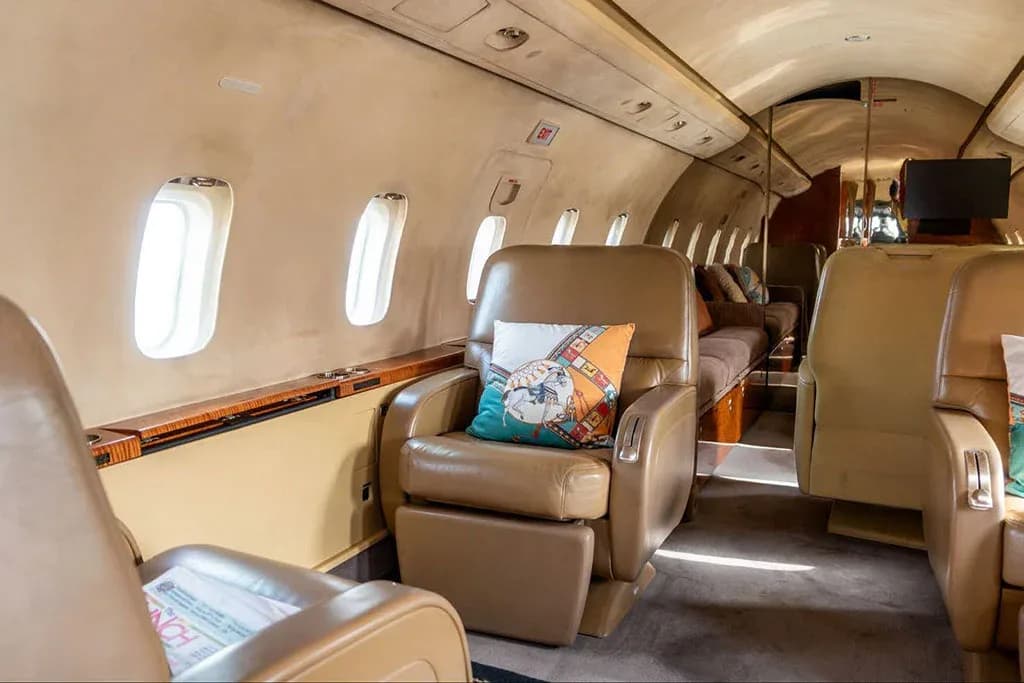 A private jet cabin interior showing tan leather seats, windows, and a lounge area in the background.