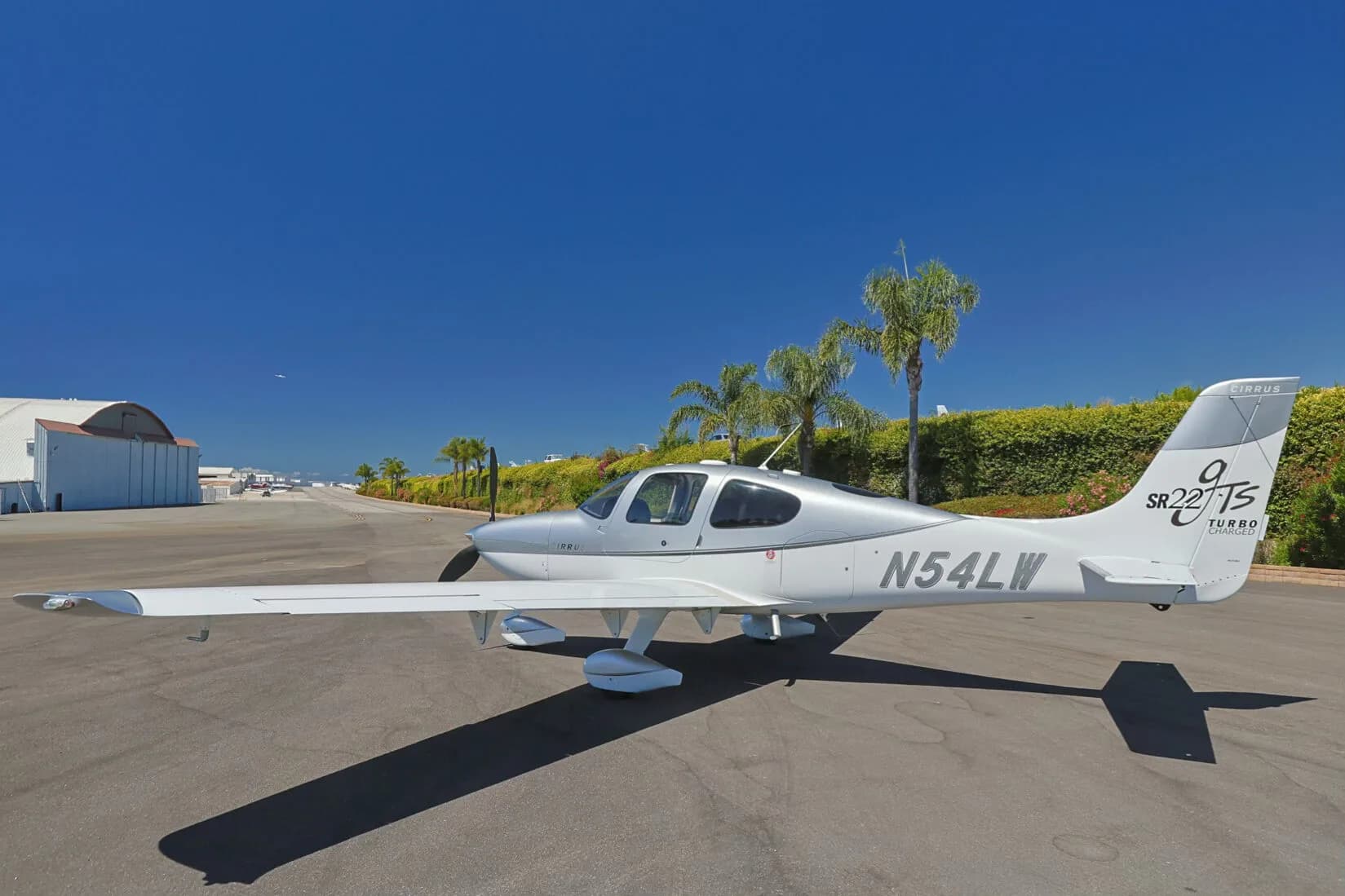Side view of a silver and white Cirrus SR22T (N54LW) on an tarmac, with hangars and palm trees in the background.
