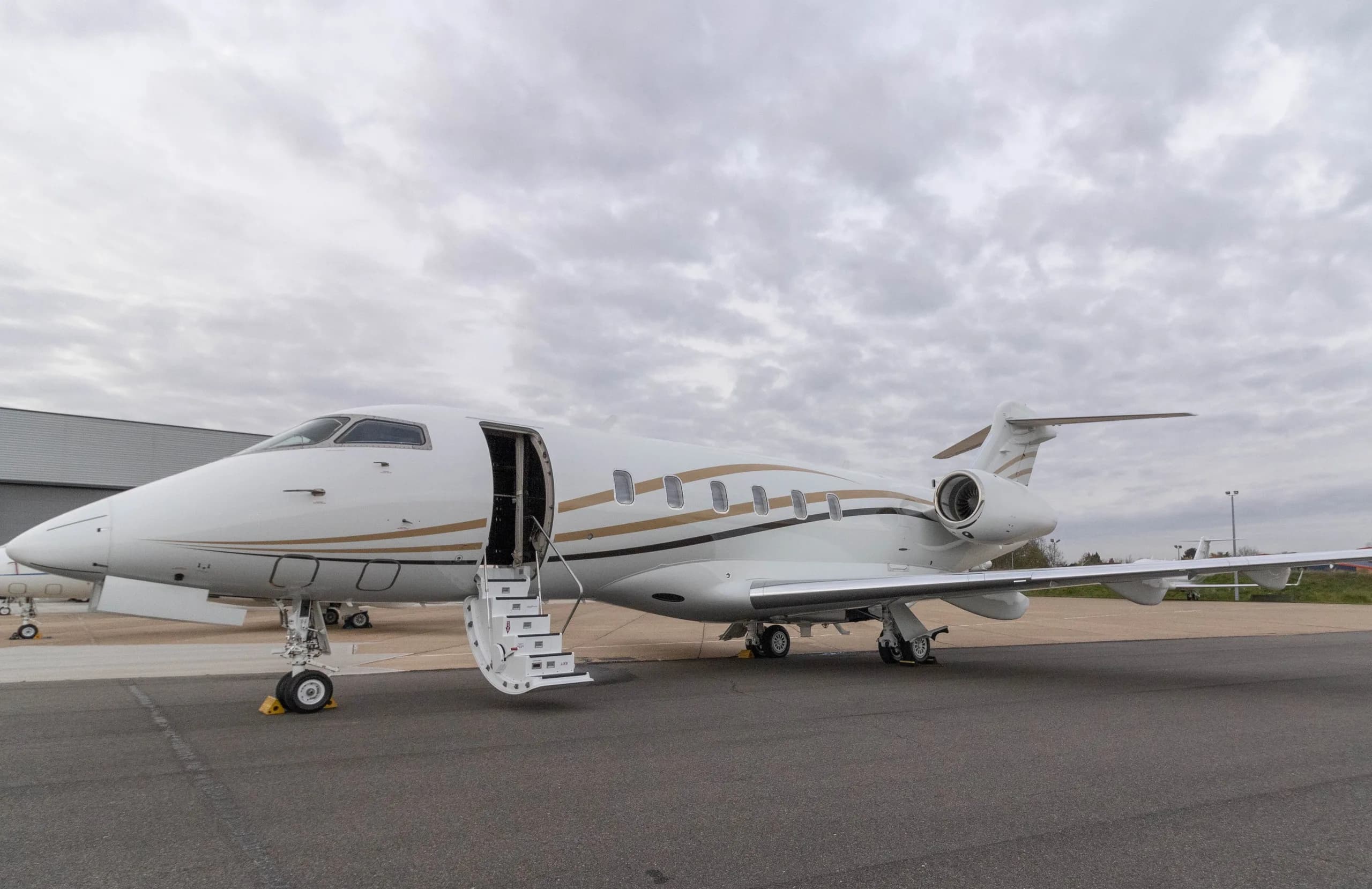 A white Bombardier Challenger 350 business jet with gold and black stripes is parked on an apron with its main door open and stairs extended under a cloudy sky.