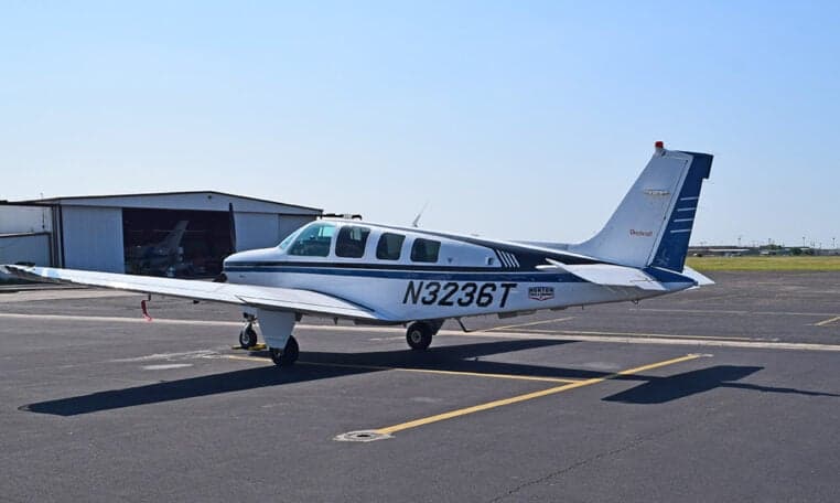 A white and blue Beechcraft Baron aircraft, N3236T, is parked on an asphalt tarmac with a hangar in the background.