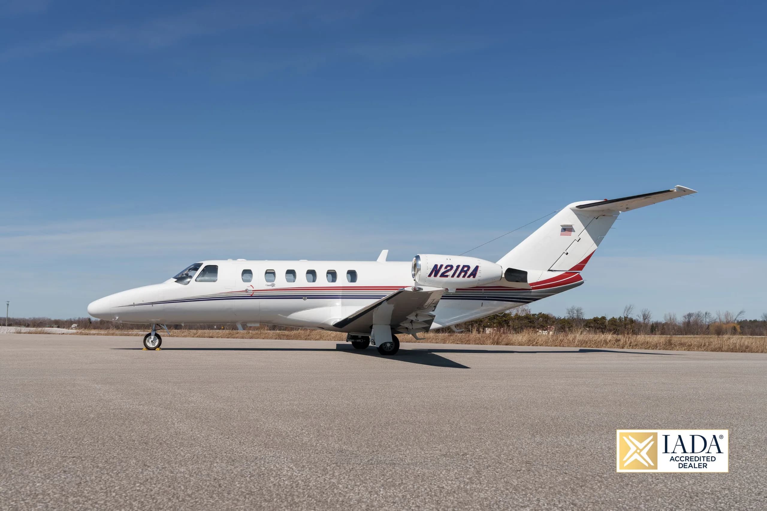 A white Cessna Citation business jet (N21RA) with red and blue stripes parked on an airfield, viewed from the side-left.