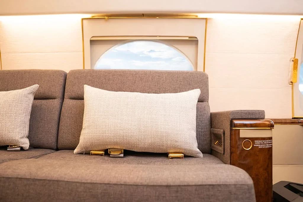 A close-up of a grey sofa with a white pillow and a window in a private jet cabin.