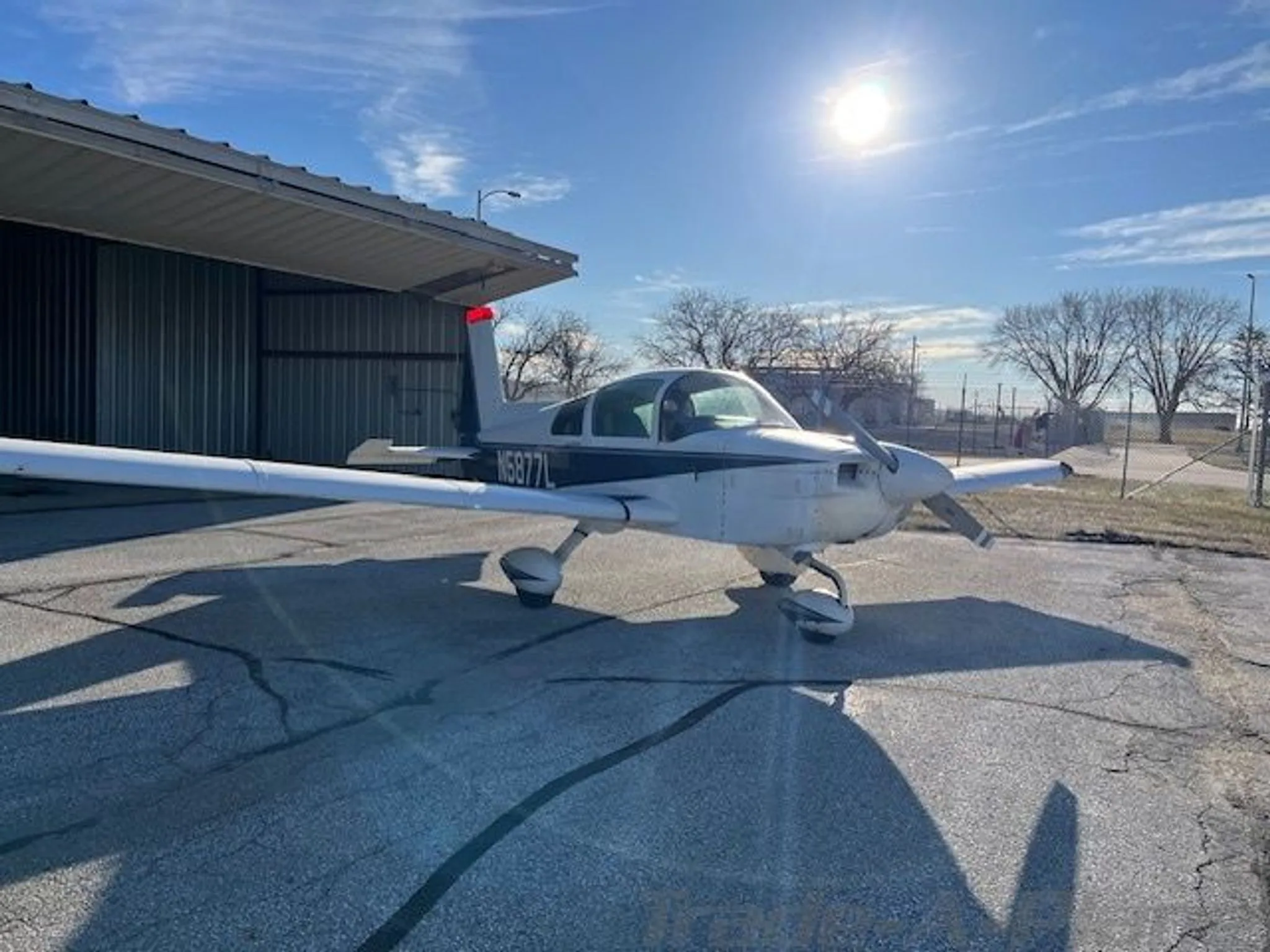 A white and blue Grumman AA-5B Tiger aircraft (N5877L) parked on a tarmac under a sunny sky.