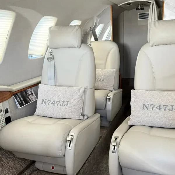 A close-up view of light-colored leather seats in a private jet cabin, with pillows embroidered with 'N747JJ'.