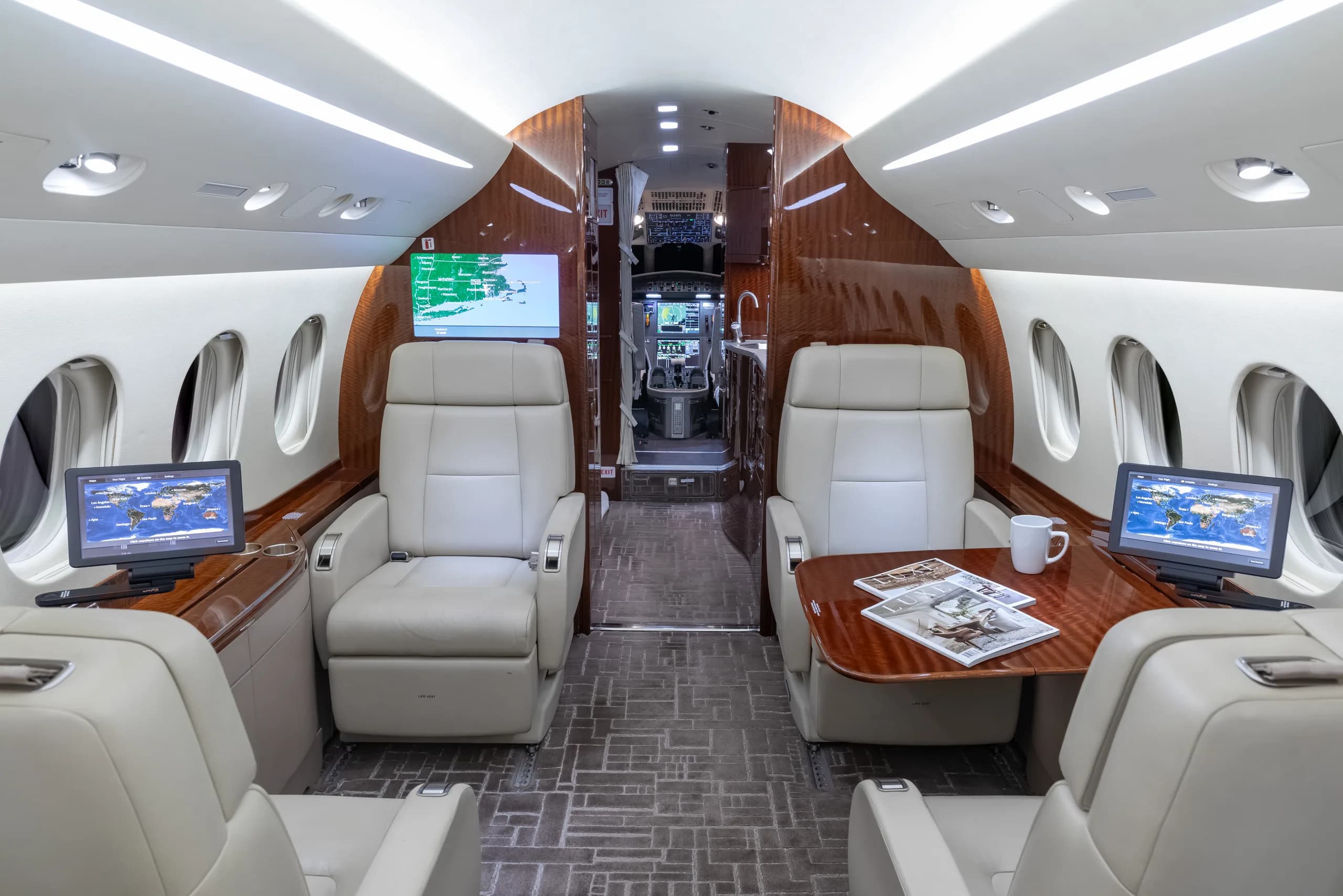 View of a luxurious aircraft cabin with light beige leather seats, two wooden tables with screens, and a glimpse of the cockpit in the background.