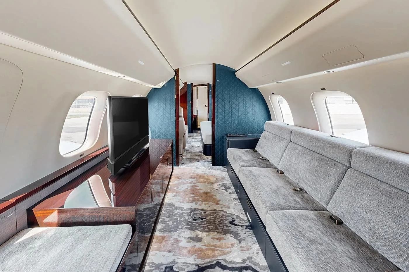 A private jet cabin lounge area featuring a long grey sofa, a large flat-screen TV, and dark wood cabinetry, with a blue patterned wall at the rear.