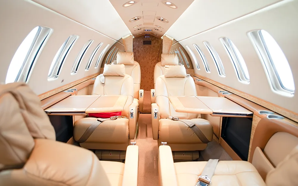 The interior of a business jet cabin with beige leather seats and fold-out tables deployed.