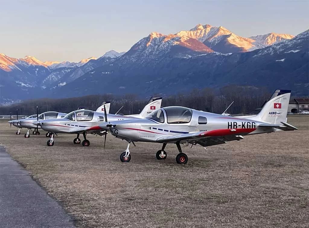 Three silver Sonaca 200 aircraft with red and blue accents parked on a grassy field with snow-capped mountains in the background at sunset.
