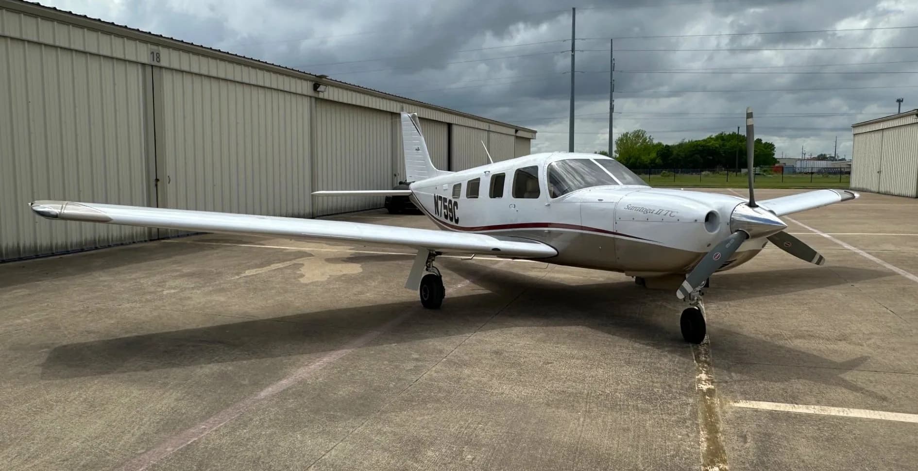 A white and tan Piper Saratoga II TC aircraft (N759C) parked on an asphalt ramp in front of hangars under a cloudy sky.