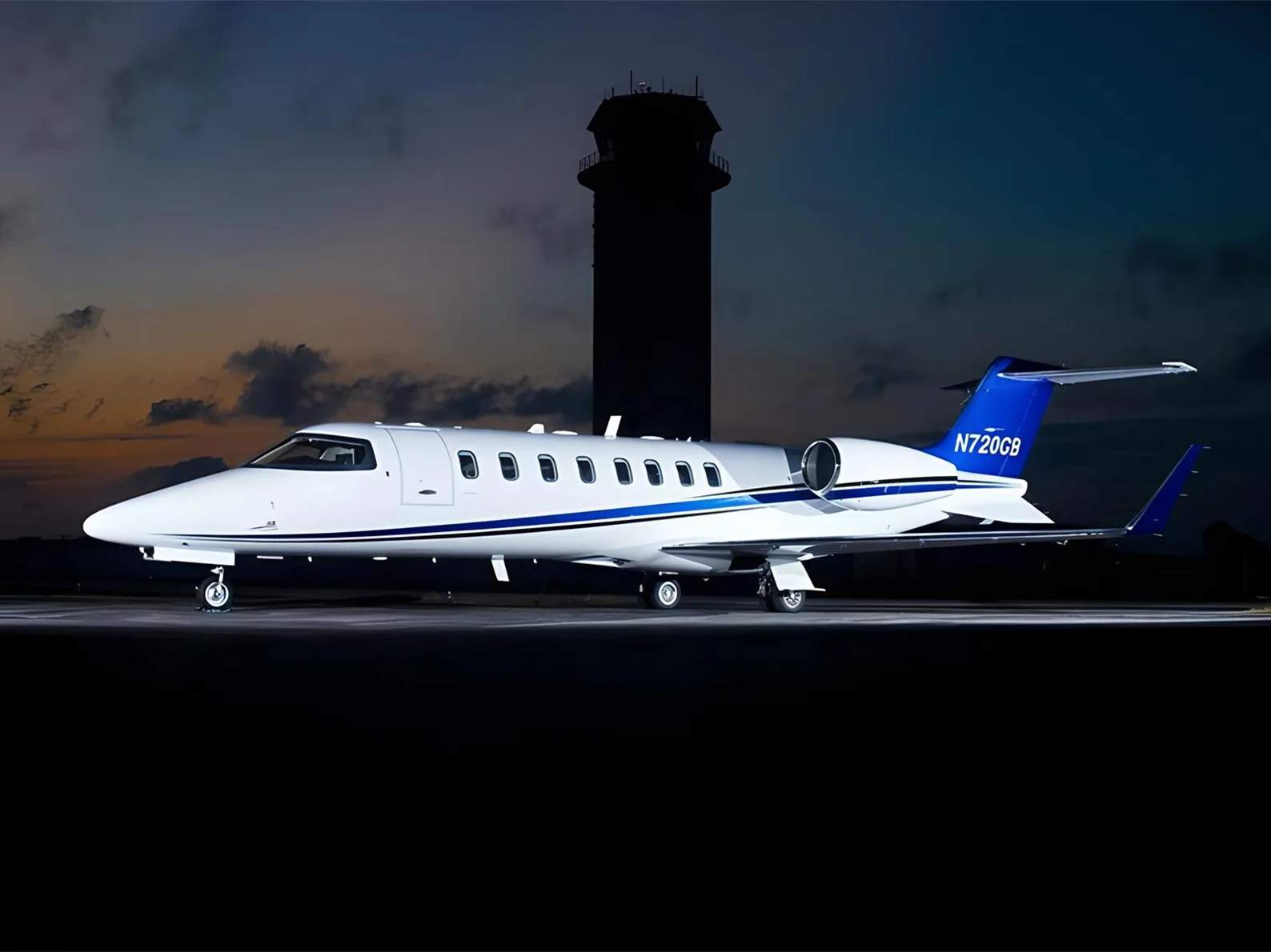A white business jet with blue stripes and tail (N720GB) parked on a tarmac at dusk with a control tower in the background.