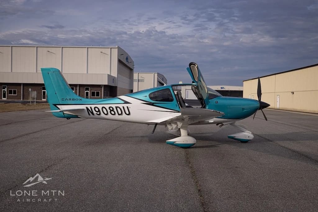A side view of the teal and white Cirrus SR22 aircraft, N908DU, with its doors open, parked on an airfield.