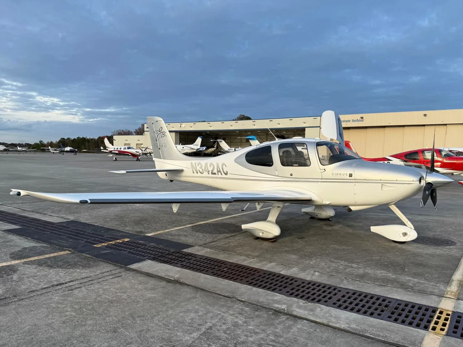 A white and silver Cirrus SR22 GTSX Turbo aircraft (N342AC) parked on an airport tarmac with hangars in the background.