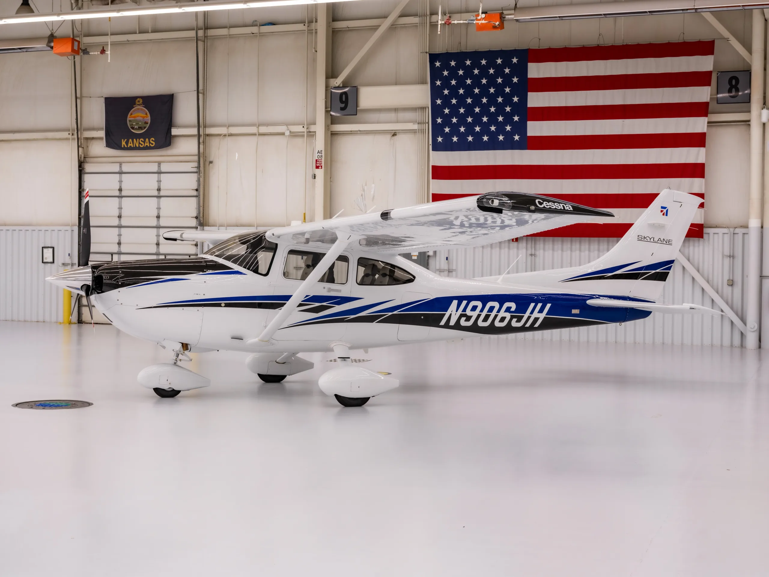 A white Cessna Skylane N906JH with blue and black accents is parked in a hangar with a US flag and Kansas flag in the background.
