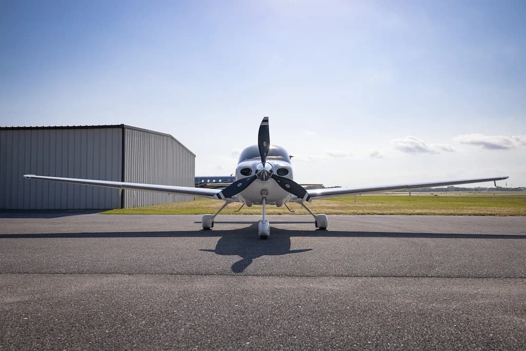 A head-on view of a white Cirrus SR22 Turbo aircraft with a three-bladed propeller, parked on an asphalt tarmac.